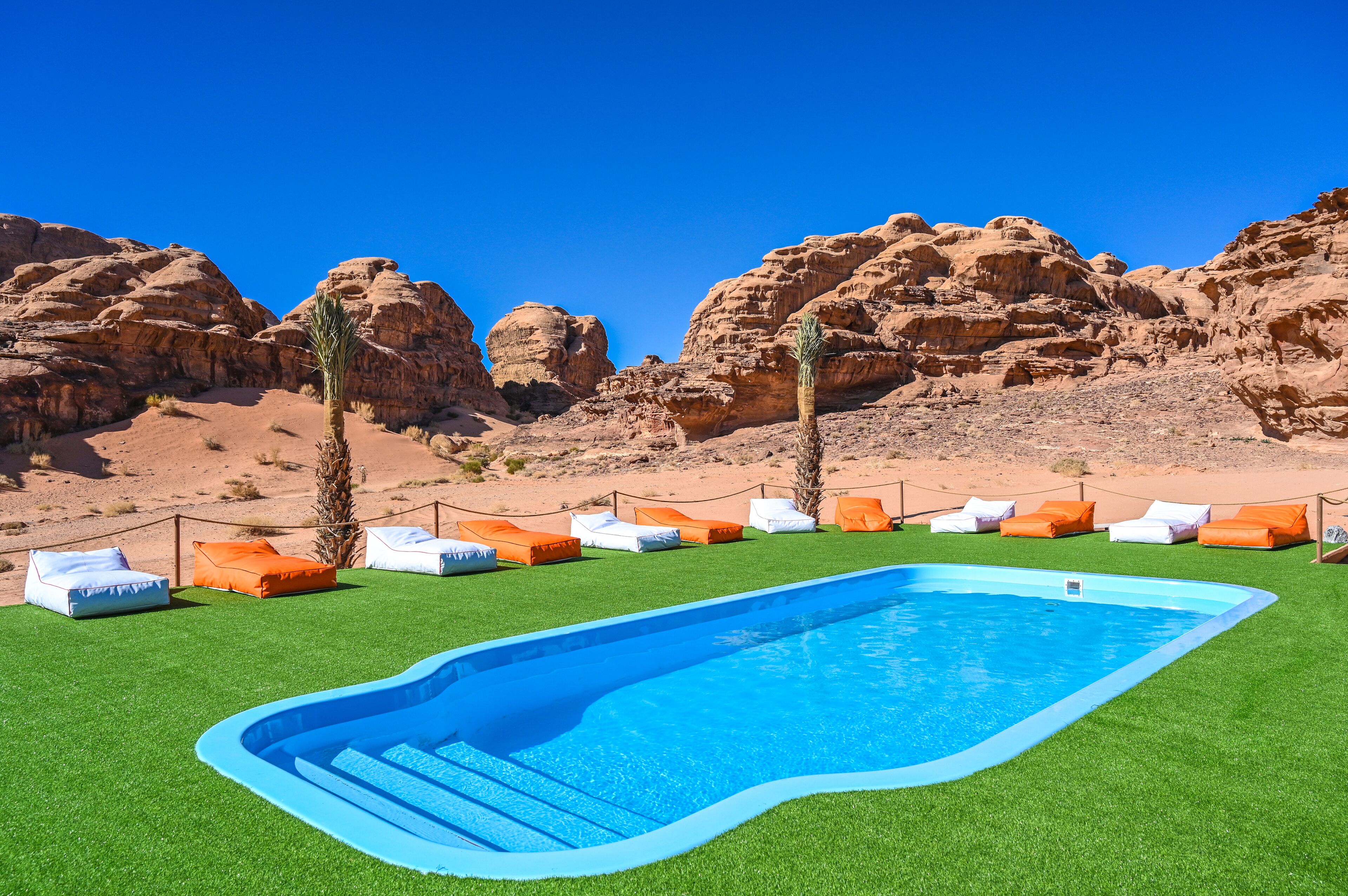 Piscina con césped artificial, rodeada de sillones de descanso y palmeras, frente a un paisaje desértico con formaciones rocosas.