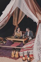Uma área de refeições coberta por tendas, com uma mesa posta para dois, rodeada por almofadas e lanternas.
