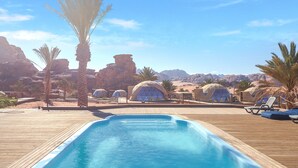 Un área de piscina con muelle de madera, palmeras y tiendas en forma de cúpula en un paisaje desértico.