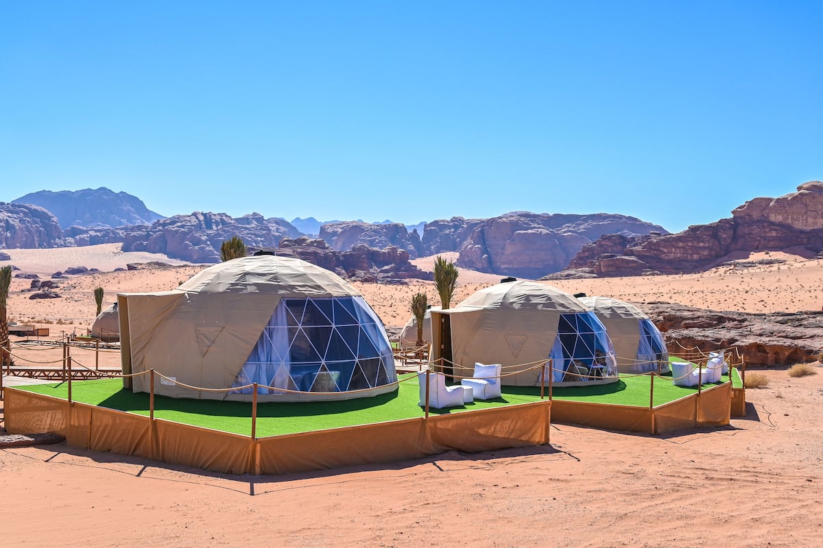Duas yurts em formato de cúpula com grama sintética verde e mobiliário branco, localizadas no deserto com montanhas rochosas ao fundo.