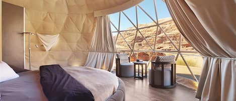 A spacious tented room with a large bed, a small table, and a view of a rocky landscape.