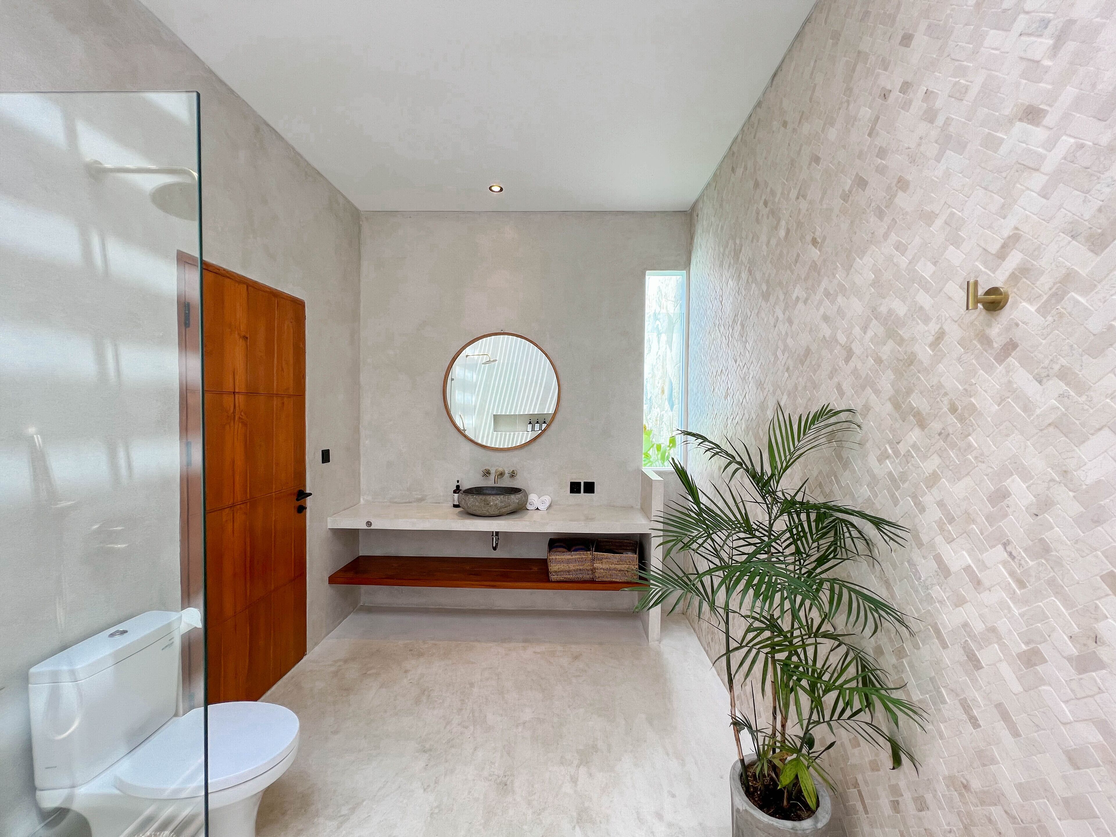 A modern bathroom with a glass shower enclosure, a toilet, a sink with a round mirror, a potted plant, and a herringbone-patterned wall.