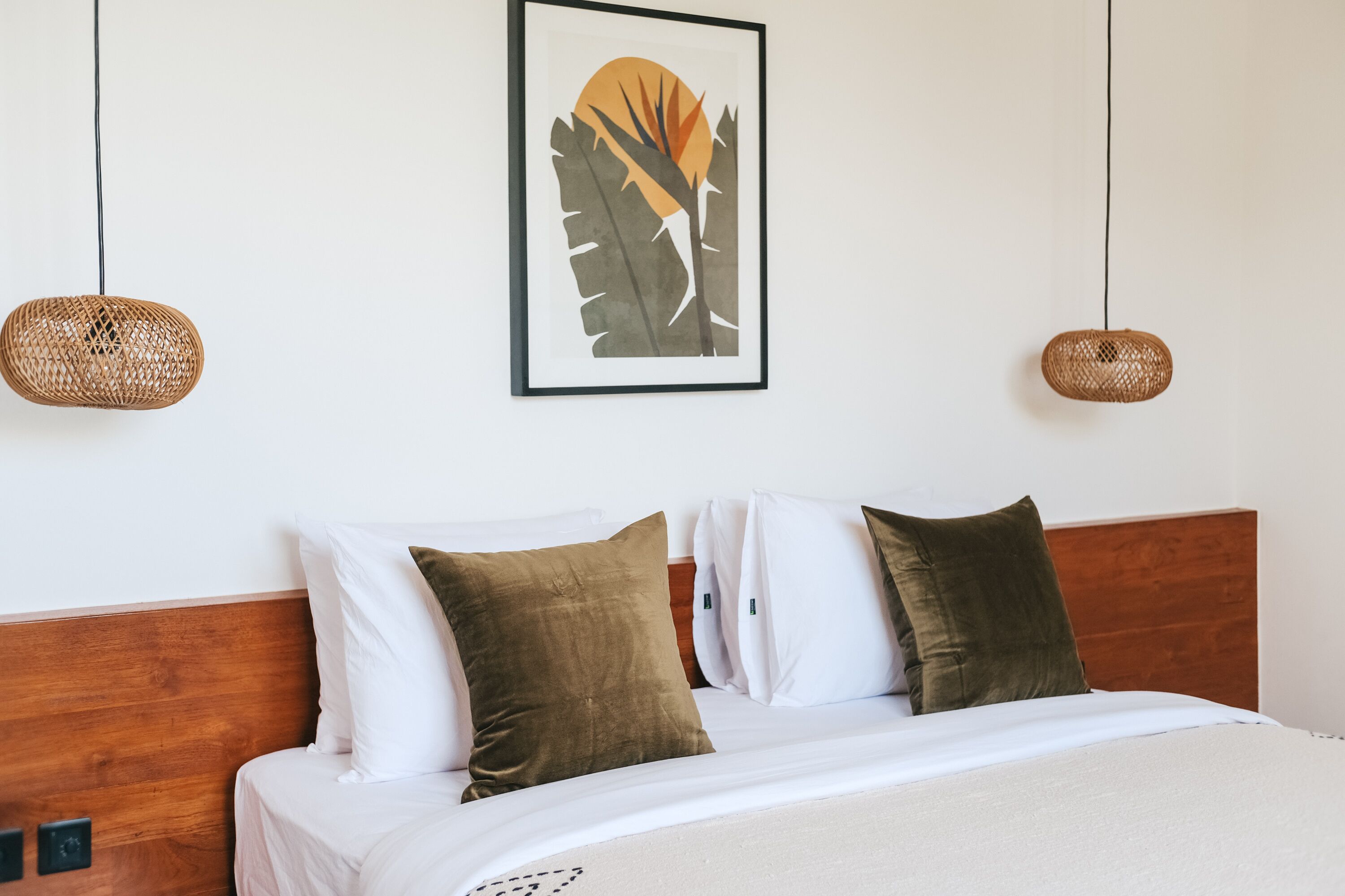 A neatly made bed with white and brown bedding, a framed artwork above it, and two woven wall hangings.
