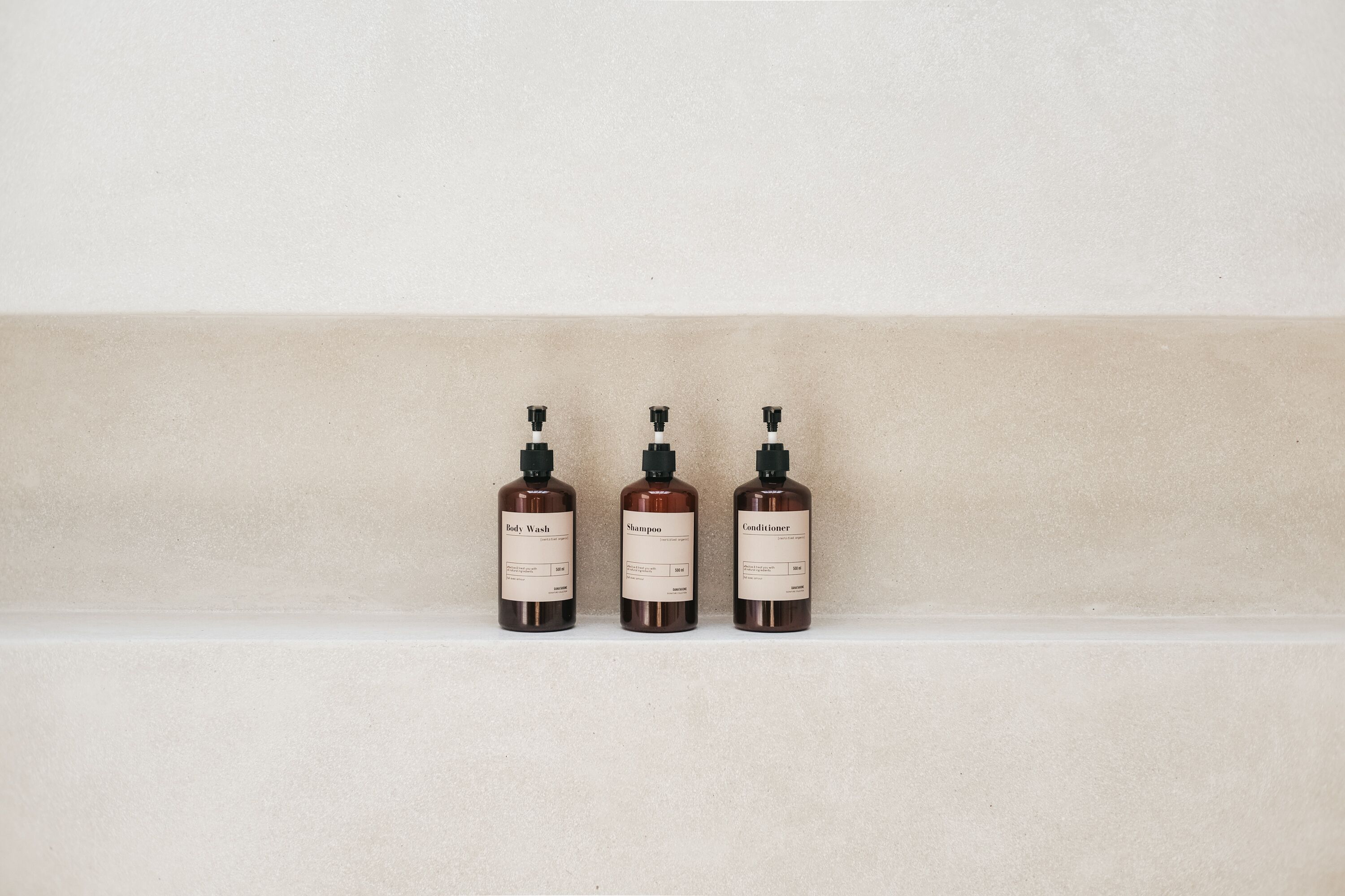 Three bottles of body wash, shampoo, and conditioner on a shelf against a plain wall.