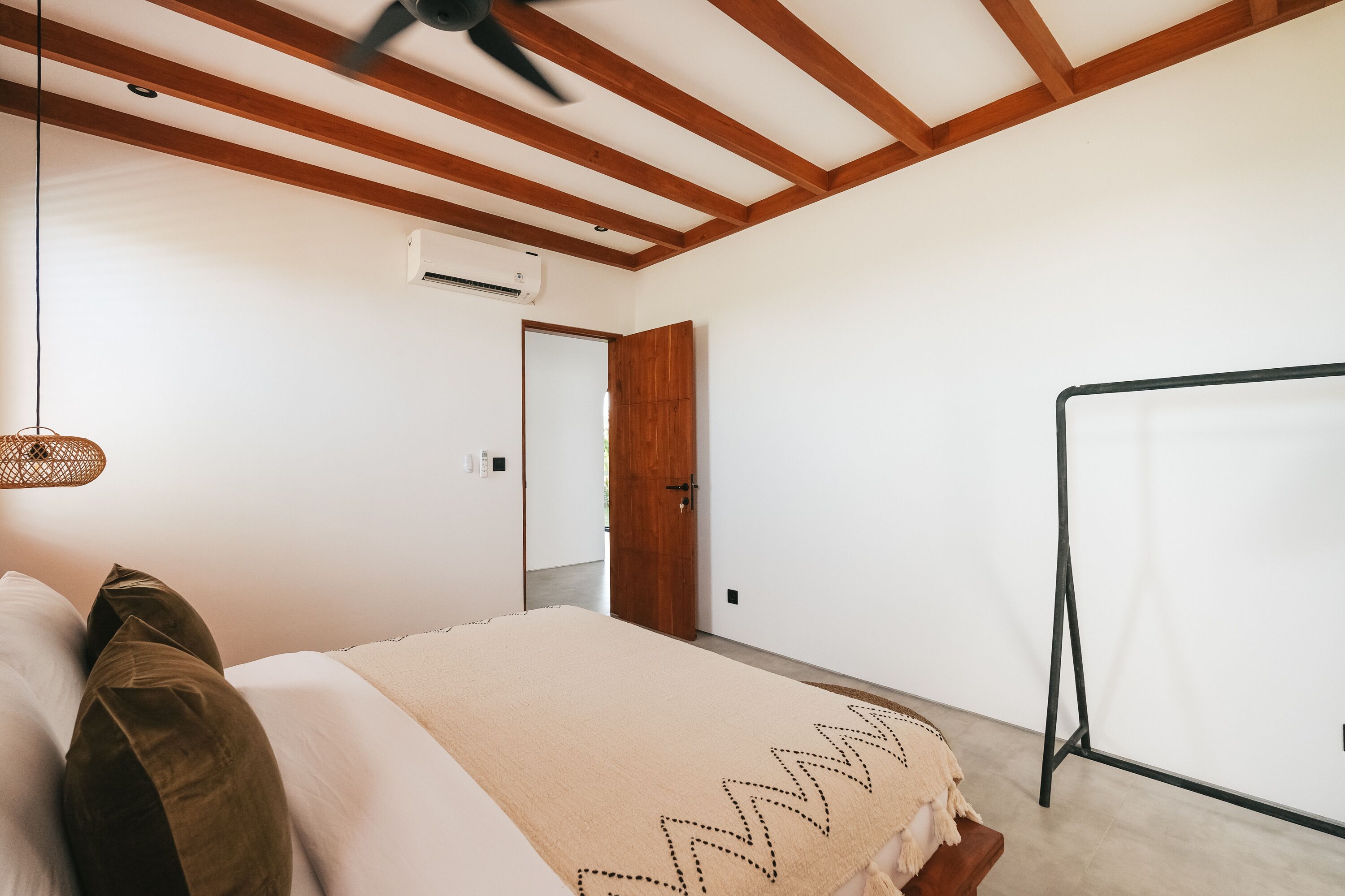 A bedroom with a bed, wooden ceiling beams, a hanging light fixture, and a door leading to another room.