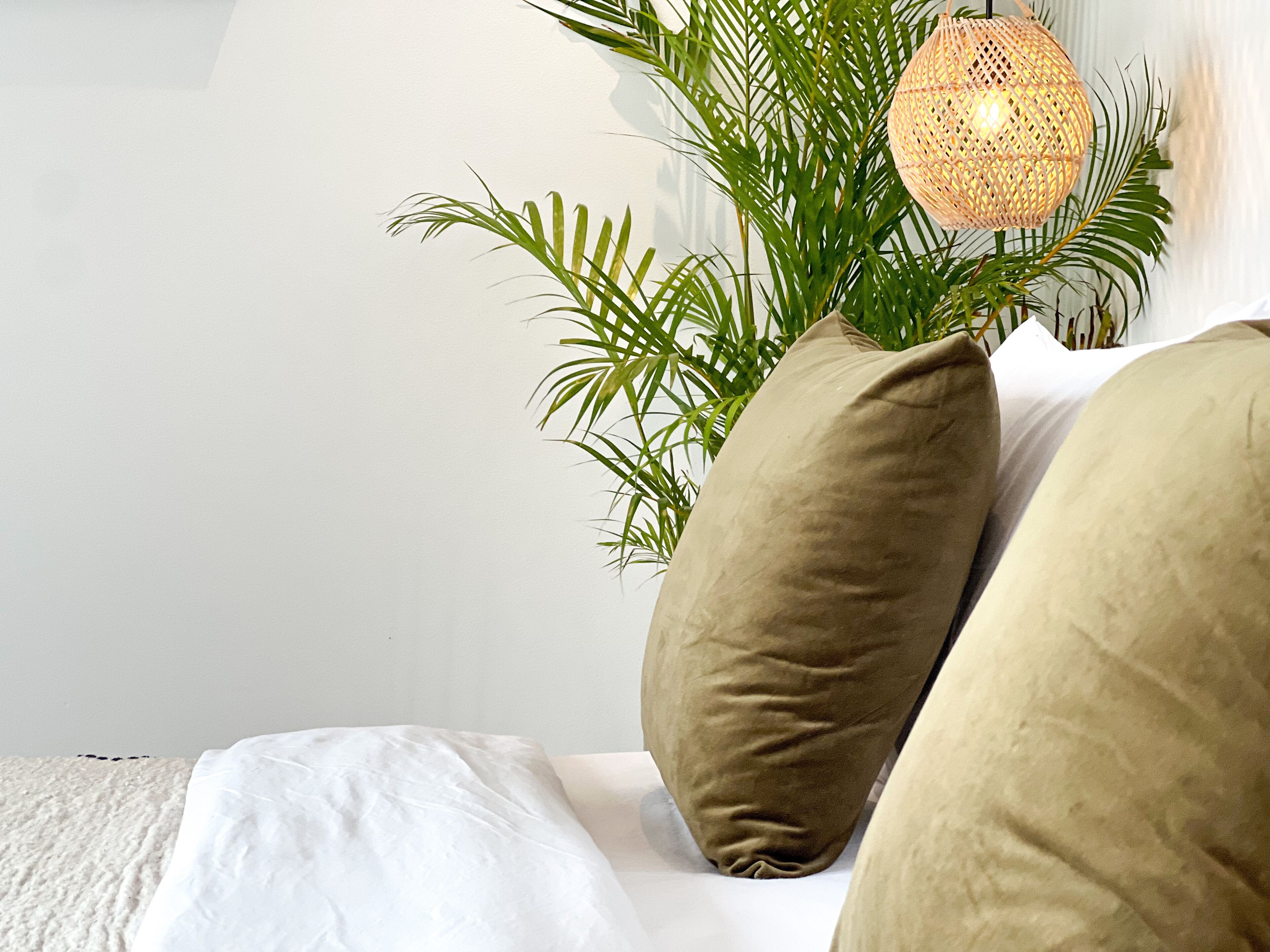 A bed with white bedding, two beige pillows, and a green plant with a woven light fixture above.