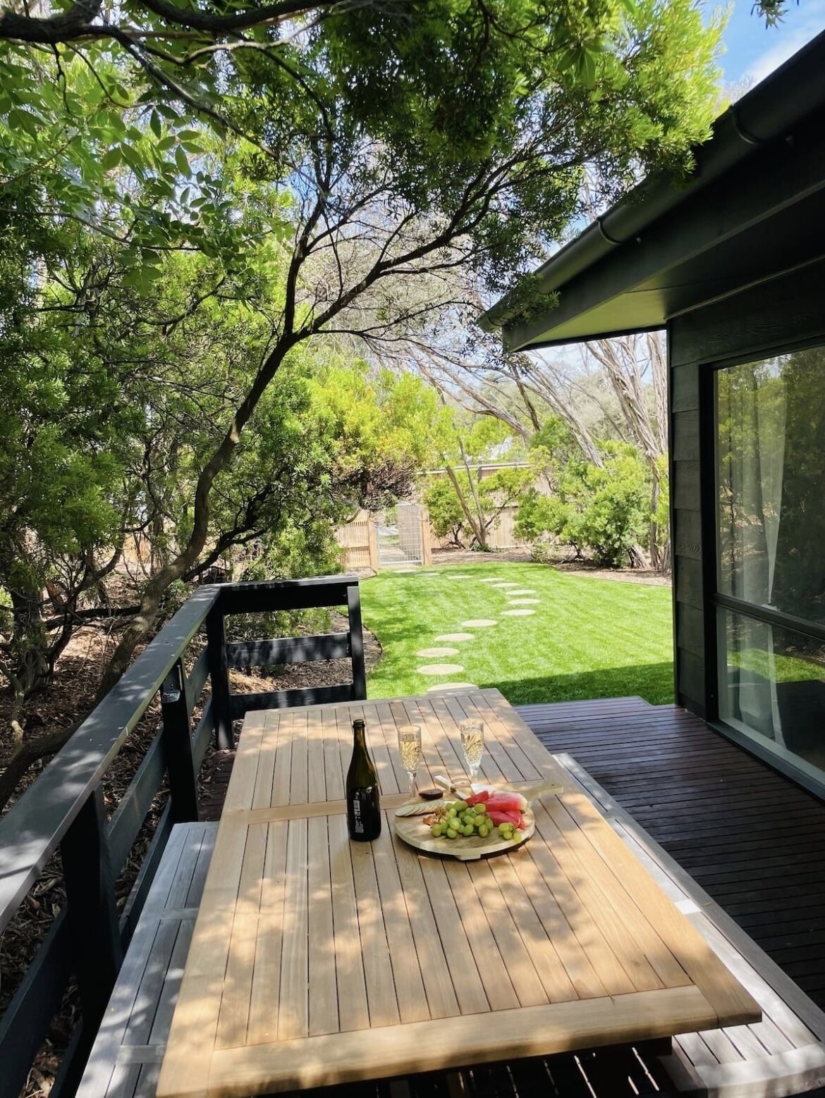 Una terraza de madera con una mesa preparada para una comida, rodeada de abundante vegetación y un césped cuidado.