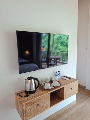 Una habitación de hotel moderna con un televisor de pantalla plana grande montado en la pared, una mesa consola de madera con una tetera, tazas y un control remoto, y vistas a un balcón con plantas y mobiliario exterior.