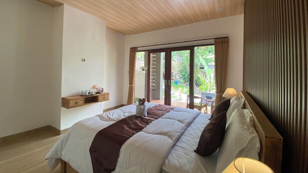 A bedroom with a large bed, a wooden ceiling, a window with curtains, and a small table with items on it. - Sulendra Jungle Suites Ubud View by EPS (Ubud)