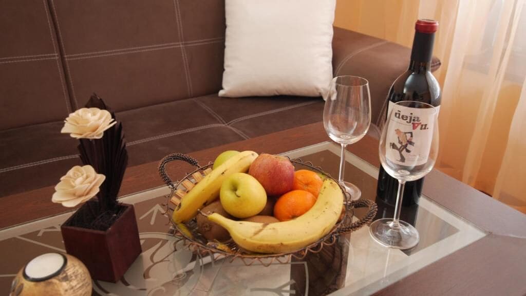 Una mesa de comedor con un frutero que contiene plátanos, manzanas y naranjas, una botella de vino y una copa.