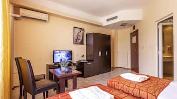 A hotel room with two beds, a desk with a television, a wardrobe, and an air conditioning unit.