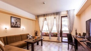 A living room with a sofa, a coffee table, a TV, and a desk with a laptop. There are curtains on the windows and a picture on the wall.