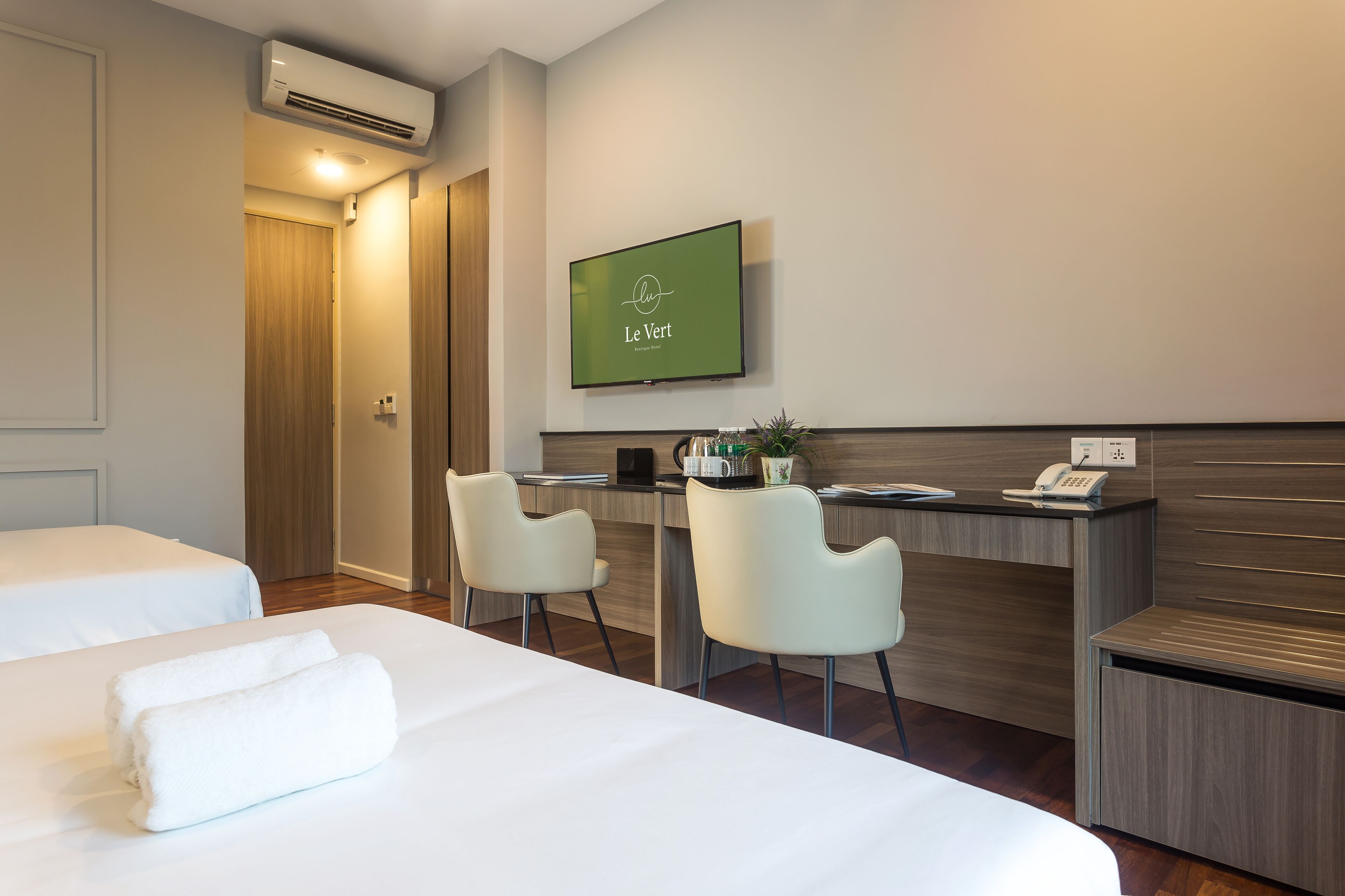 A hotel room with two beds, a desk with chairs, a TV, and an air conditioning unit.
