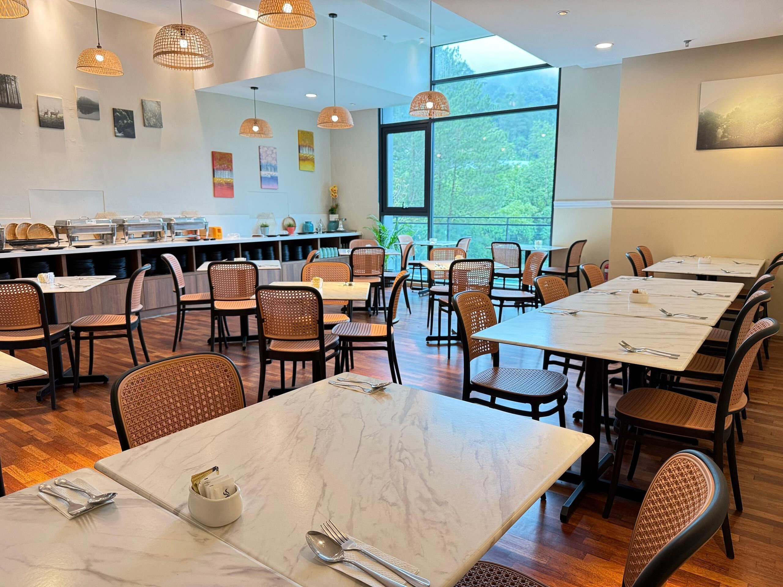 A spacious dining area with multiple tables and chairs, large windows offering a view of greenery, and a buffet setup with various food items.