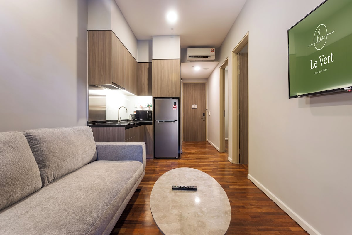 Une chambre d’hôtel moderne avec un canapé gris, une table basse ronde, un coin cuisine et un téléviseur fixé au mur qui diffuse des images de « Le Vert Boutique Hotel ».