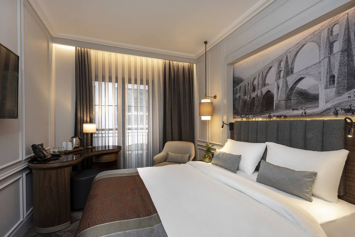 A hotel room with a large bed, a desk with a telephone, a chair, and a painting of a bridge on the wall.