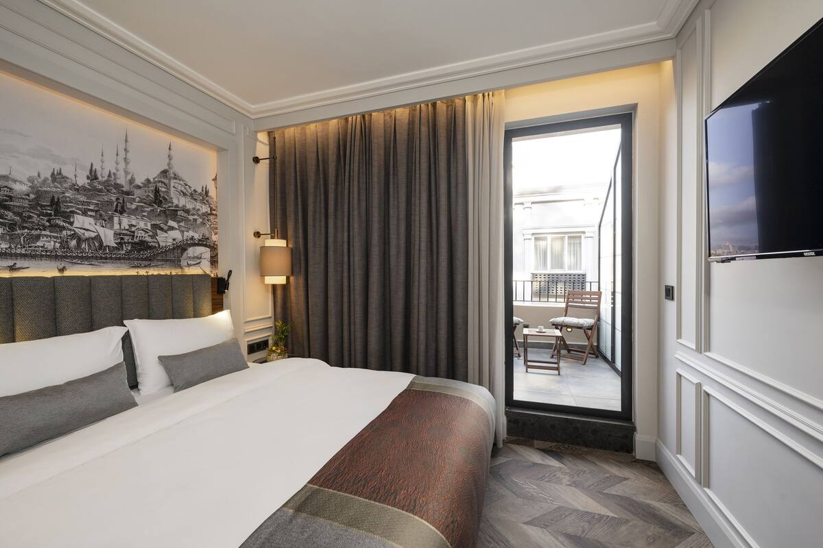 A hotel room with a large bed, a painting on the wall, a television, and a view of the city through the window.
