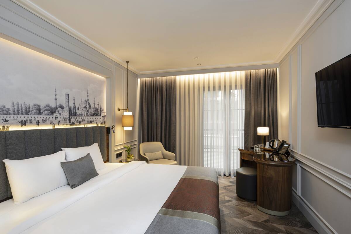 A hotel room with a large bed, a desk with a lamp, a chair, and a TV on the wall.