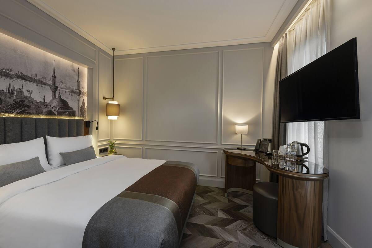 A hotel room with a large bed, a desk with a flat-screen TV, a lamp, and a painting of a cityscape on the wall.