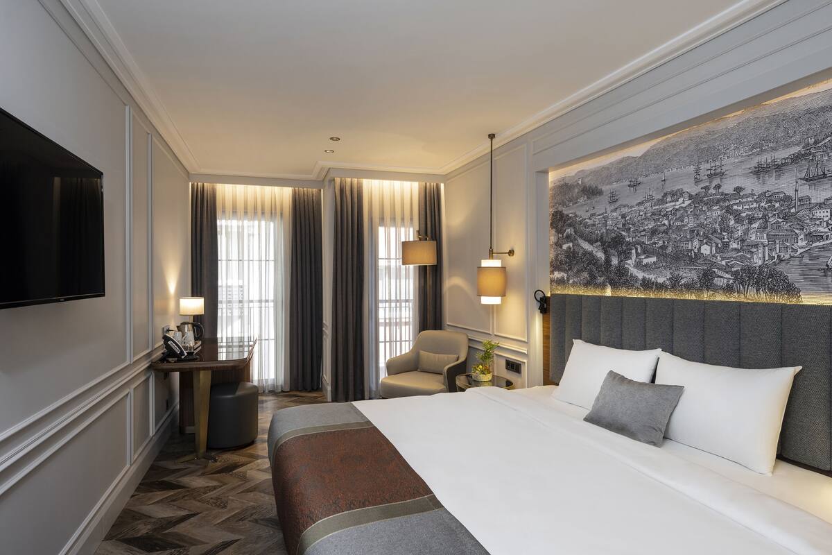 A hotel room with a large bed, a desk with a lamp, a chair, and a wall-mounted TV.