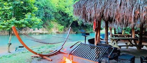Una hamaca, un pozo de fuego y una estructura de techo de paja junto a un río.