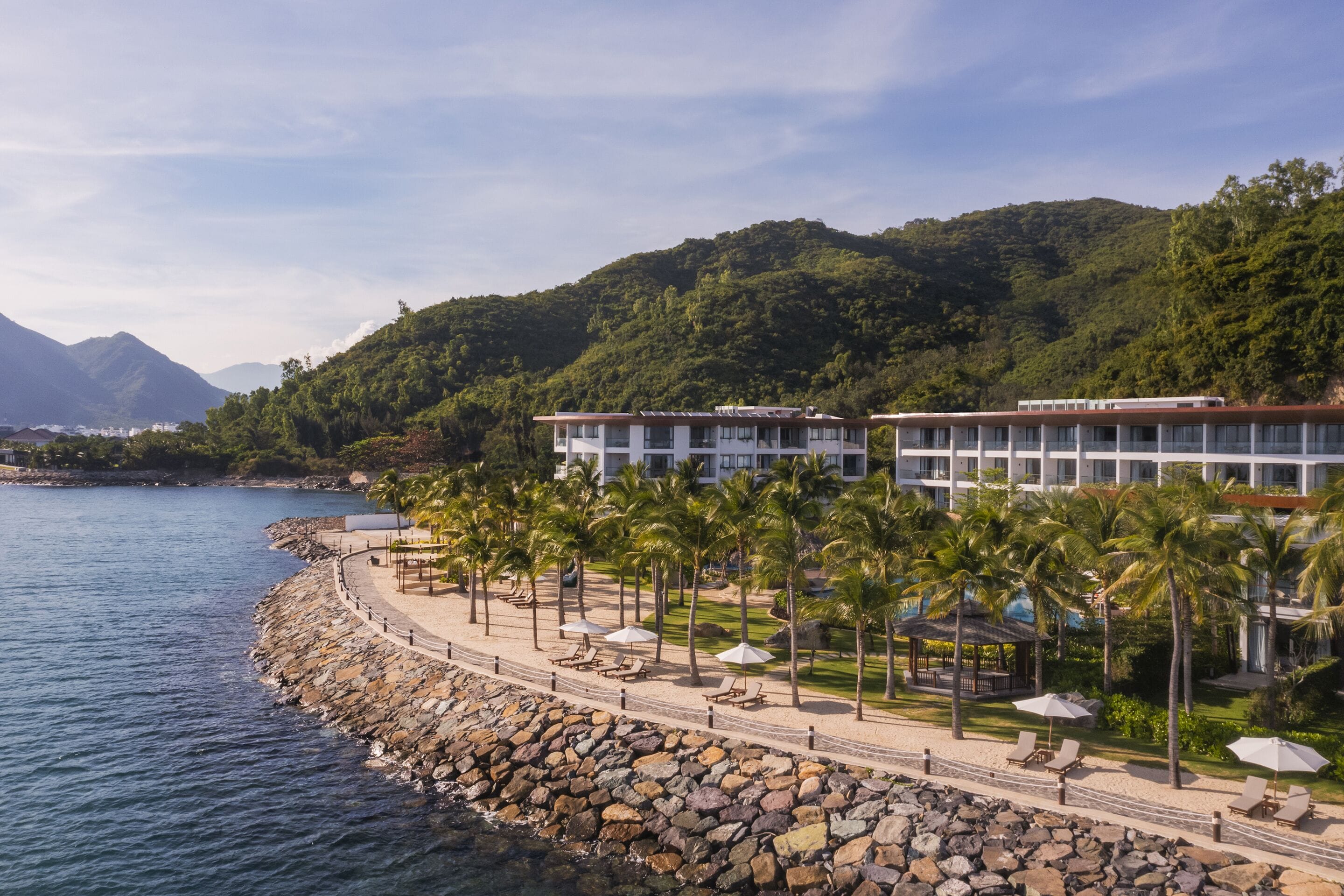Ein Hotel direkt am Strand mit felsiger Küste, Palmen und einem Berg im Hintergrund.
