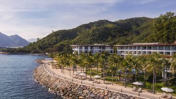 Ein Hotel direkt am Strand mit felsiger Küste, Palmen und einem Berg im Hintergrund.