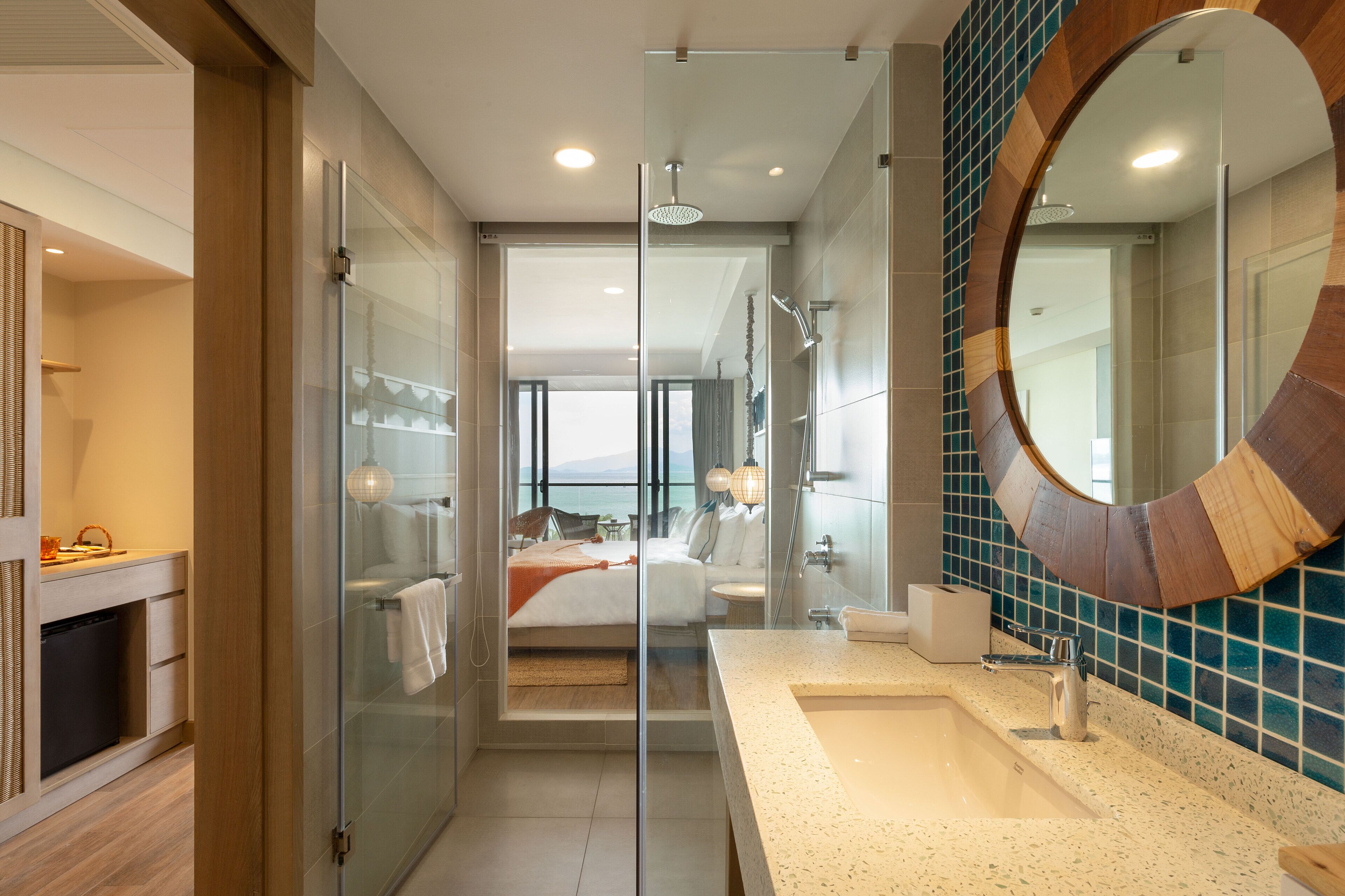 A modern bathroom with a glass shower enclosure, a large mirror, and a view of a bedroom with a bed and a balcony.