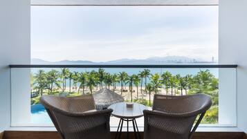 A balcony with wicker chairs and a table, overlooking a beach with palm trees and a city skyline.