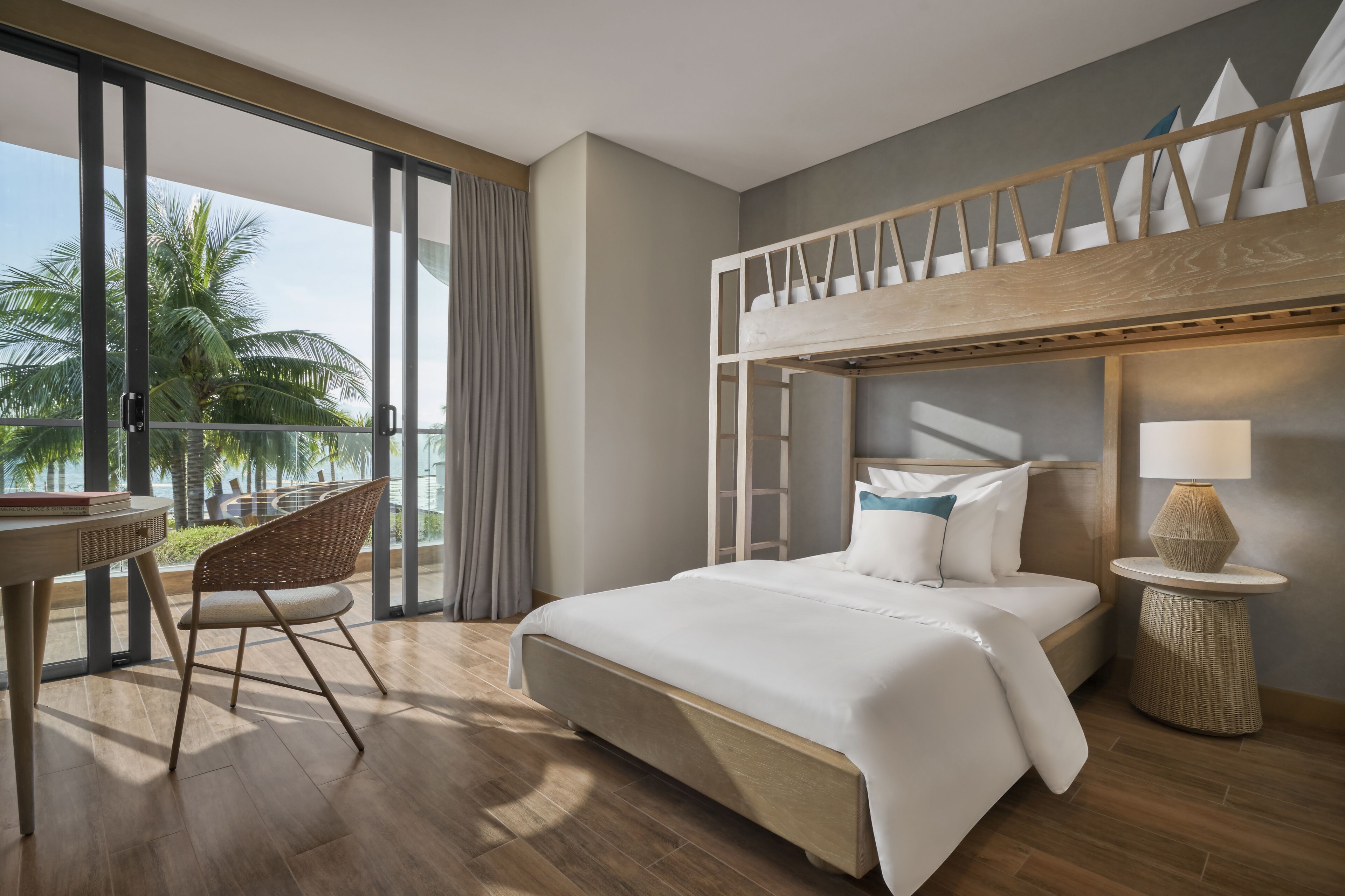 A modern hotel room with a bunk bed, a balcony with a view of palm trees, a round dining table, and a wicker side table.