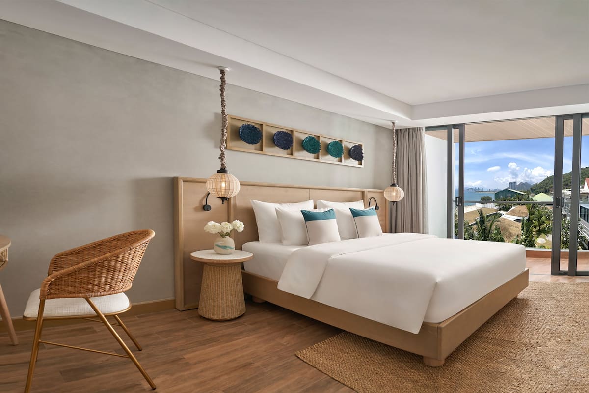 Une chambre d’hôtel moderne avec un grand lit, un fauteuil en osier, une petite table d’appoint avec une lampe, et une vue sur la mer.