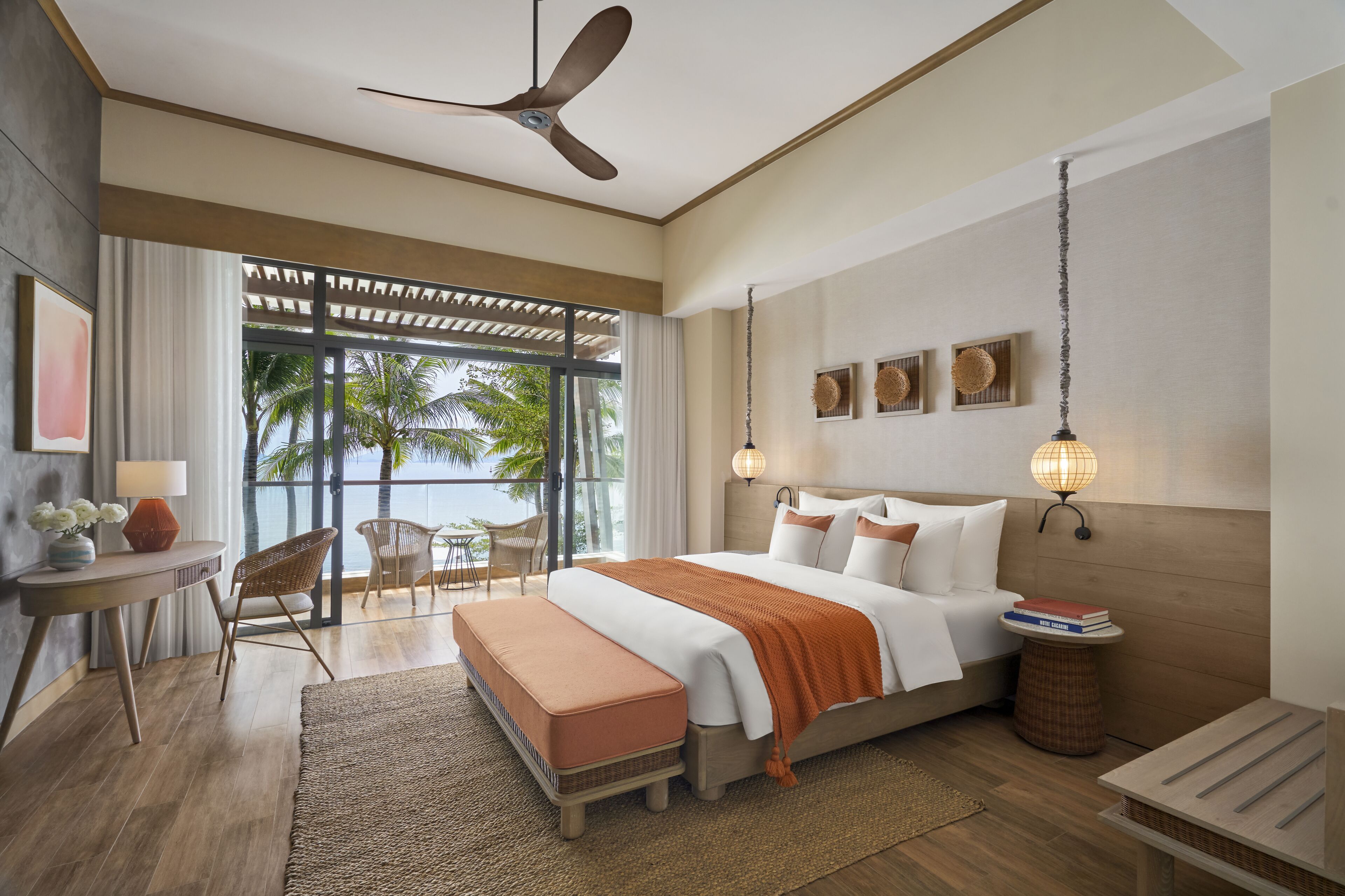 A modern hotel room with a large bed, a ceiling fan, a balcony with a view of palm trees, and a desk with a lamp.