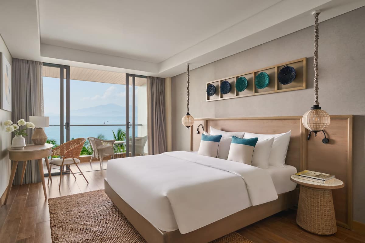 Une chambre d’hôtel moderne avec un grand lit, un balcon donnant sur la mer et une petite table avec une lampe.
