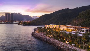 Un hotel frente al mar con paseo marítimo, palmeras y montañas al fondo.