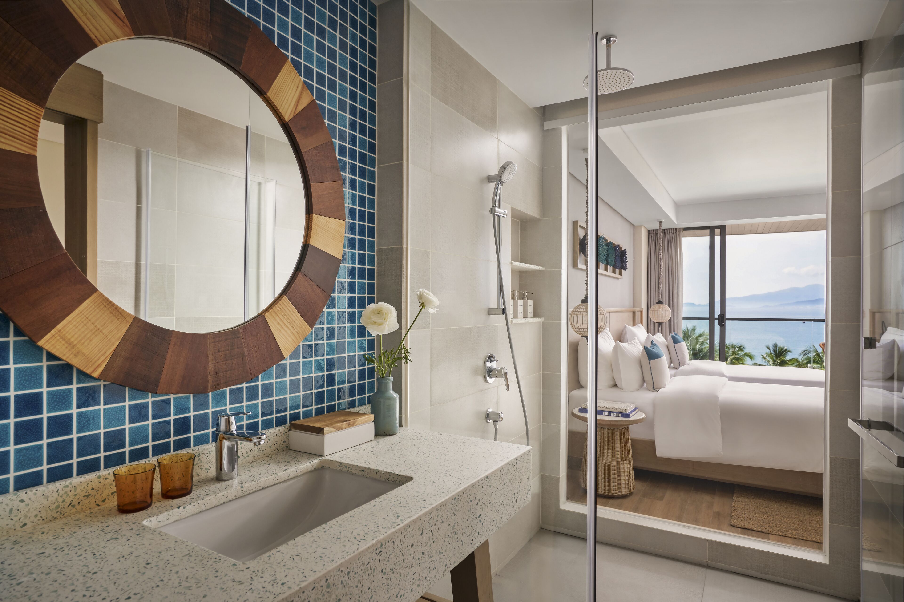 A modern bathroom with a large mirror, a sink, and a shower. There is a view of a bedroom with a bed and a balcony.