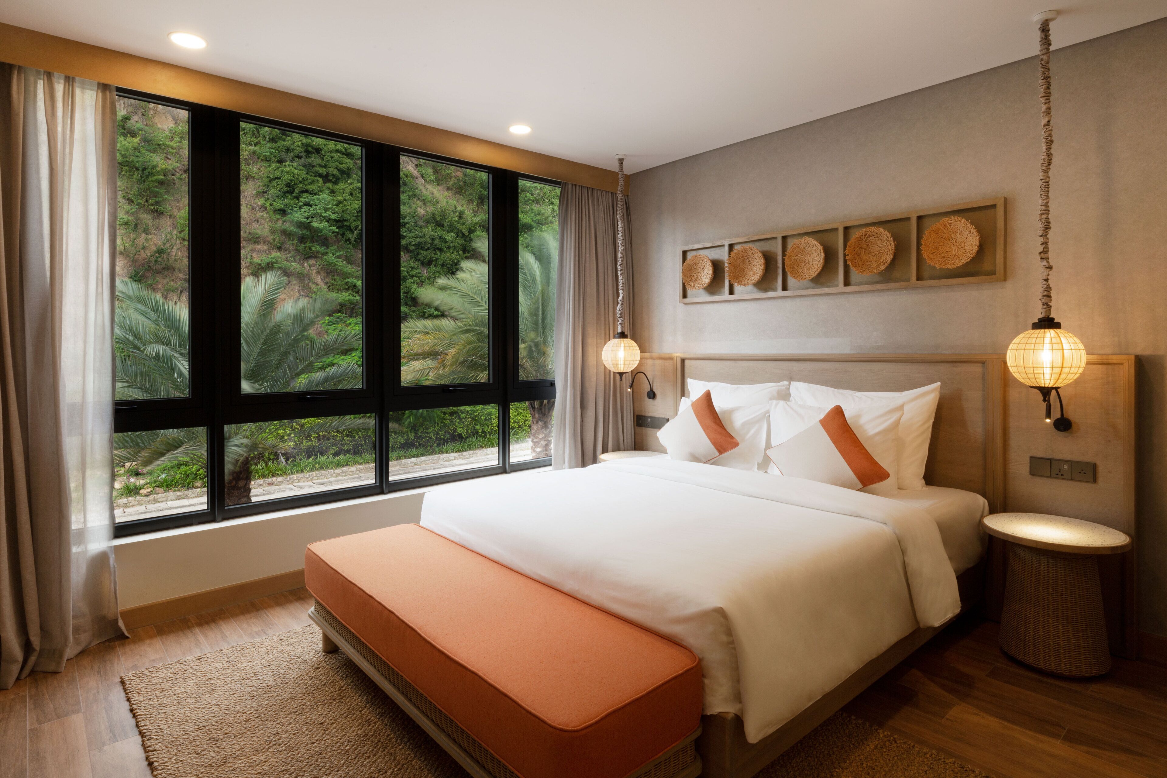A modern bedroom with a large bed, a bench, a bedside table, and a view of greenery through large windows.