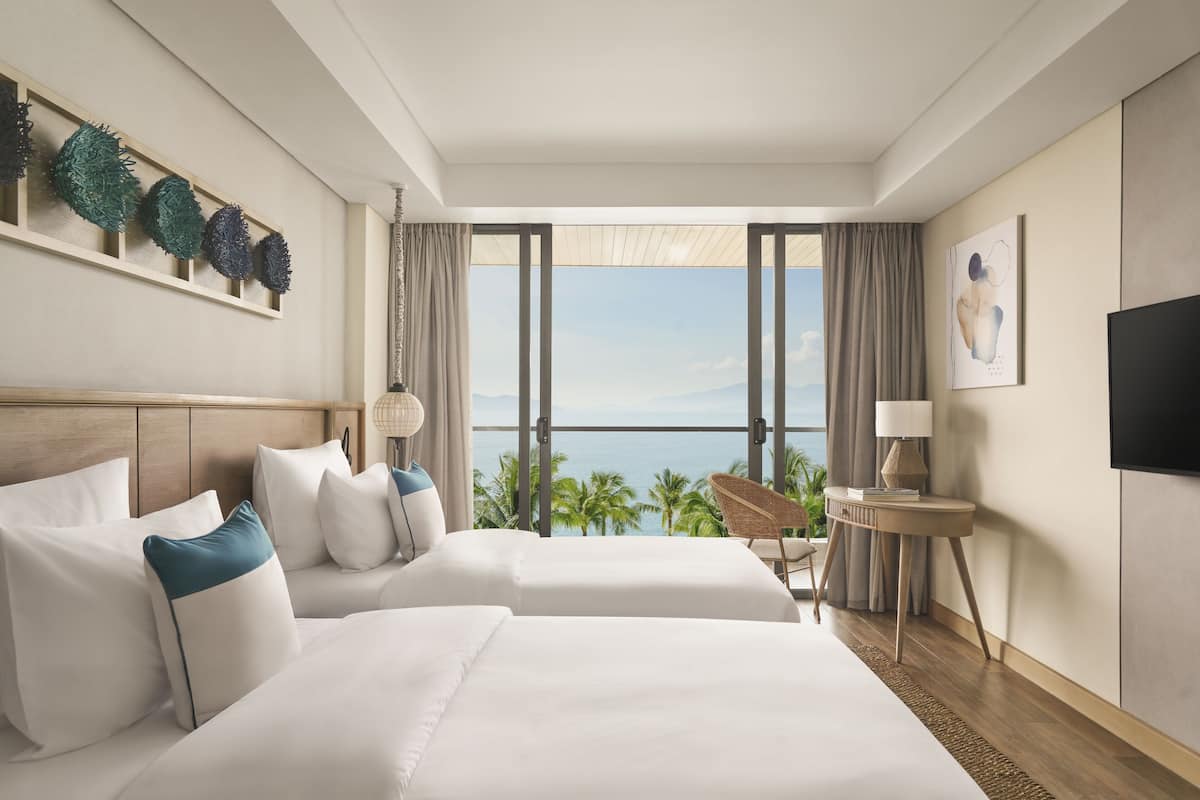 Une chambre d’hôtel avec deux lits, une grande fenêtre donnant sur des palmiers, un téléviseur à écran plat et une petite table avec une lampe.