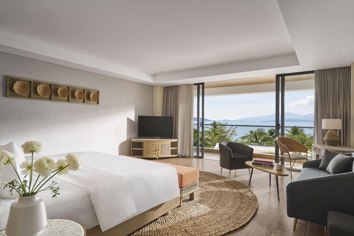 Une chambre d’hôtel moderne dotée d’un grand lit, d’une télévision, d’un coin salon avec vue sur la mer et d’un balcon offrant une vue sur les montagnes.