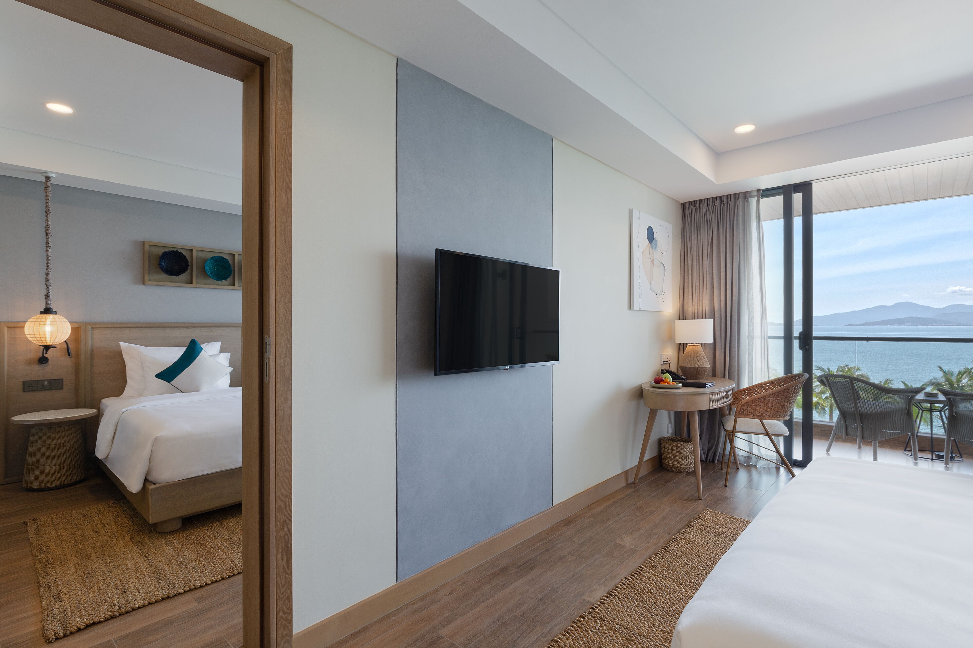 A modern hotel room with a bed, a flat-screen TV, a small table with chairs, and a balcony with a view of the sea.