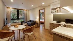 Una habitación de hotel moderna con zona de comedor, un sofá, una mesita con fruta y una cocina, además de vistas al exterior.