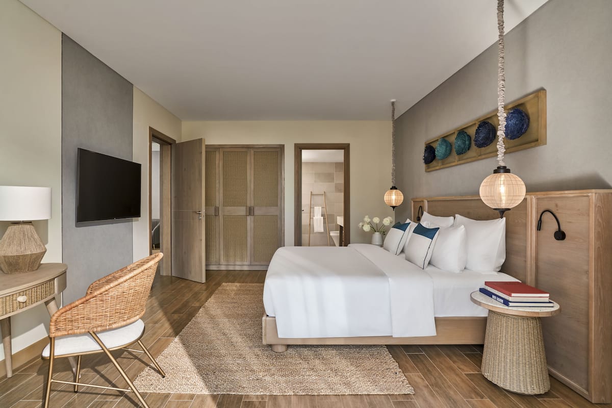 Une chambre d’hôtel moderne avec un grand lit, une télévision, un fauteuil en osier et une petite table d’appoint avec des livres.