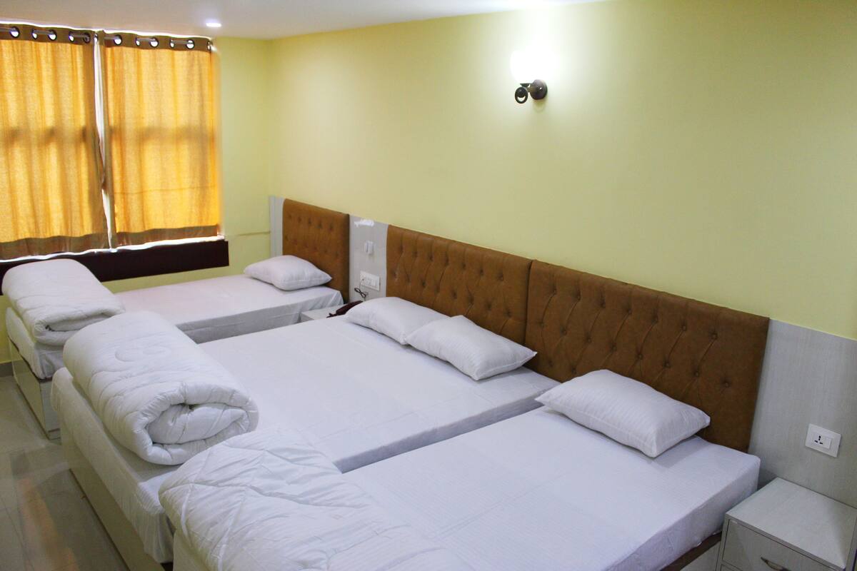 Habitación de hotel con tres camas, cada una con ropa y almohadas blancas, cabeceros de madera y una mesita de noche pequeña con un cajón.