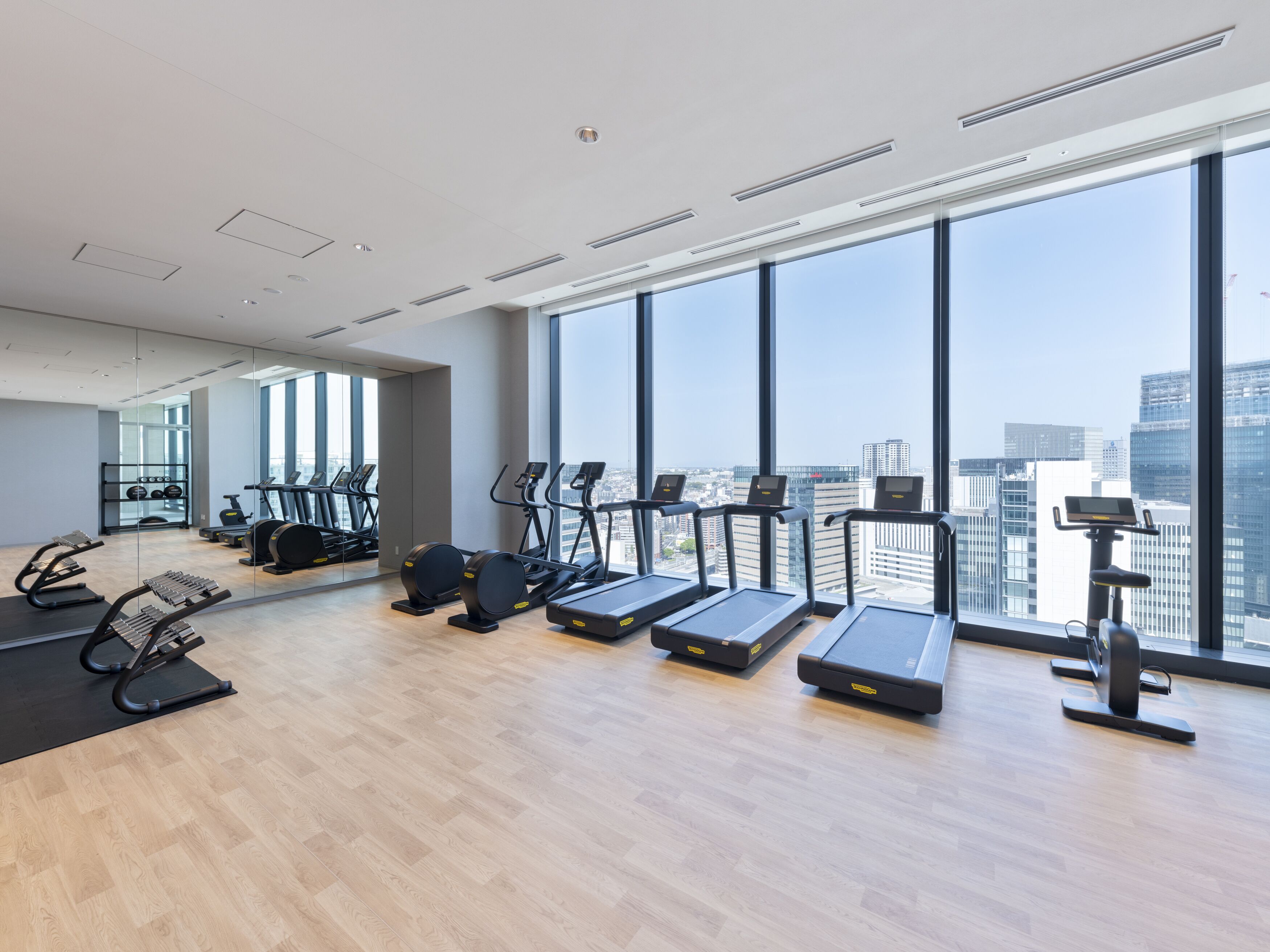 A modern gym with treadmills, elliptical machines, and a rowing machine, large windows with a city view, and a wooden floor.