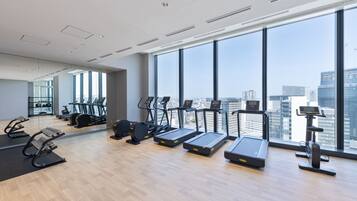 A modern gym with treadmills, elliptical machines, and a rowing machine, large windows with a city view, and a wooden floor.