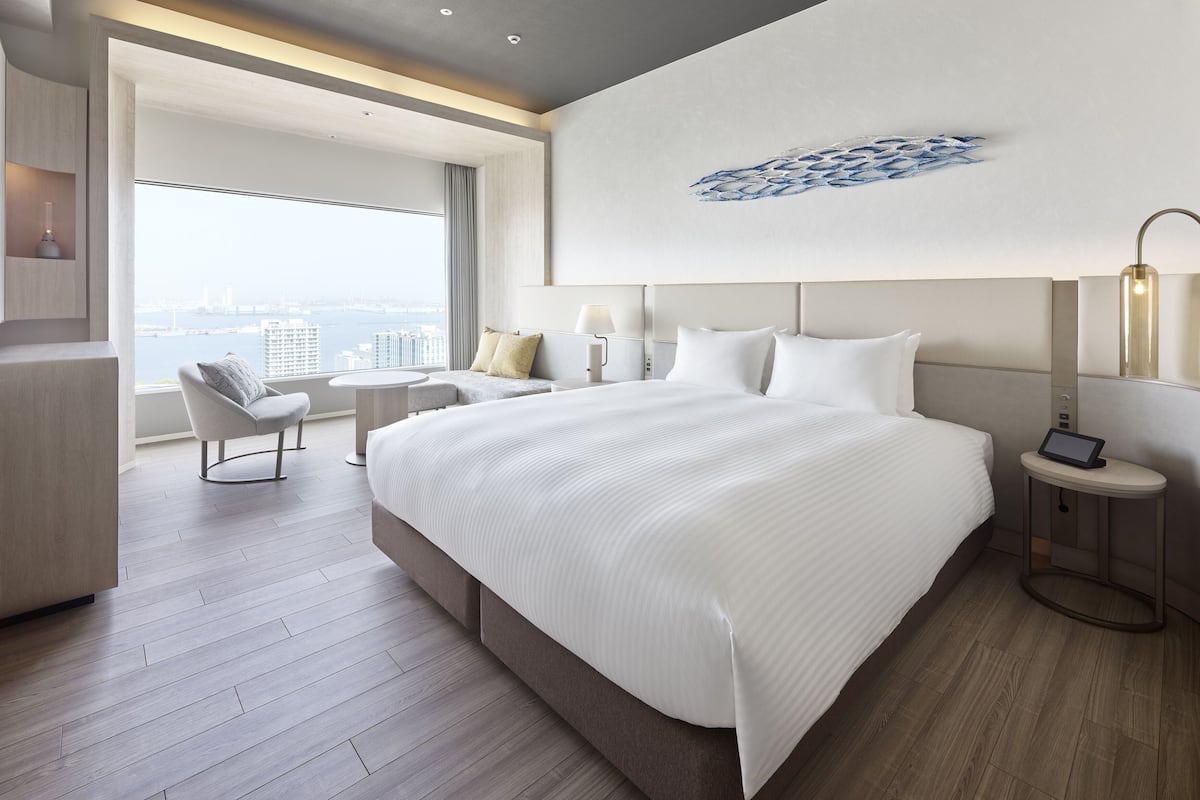 A modern hotel room with a large bed, a seating area with a sofa and armchairs, a bedside table with a lamp, and a view of the city through large windows.