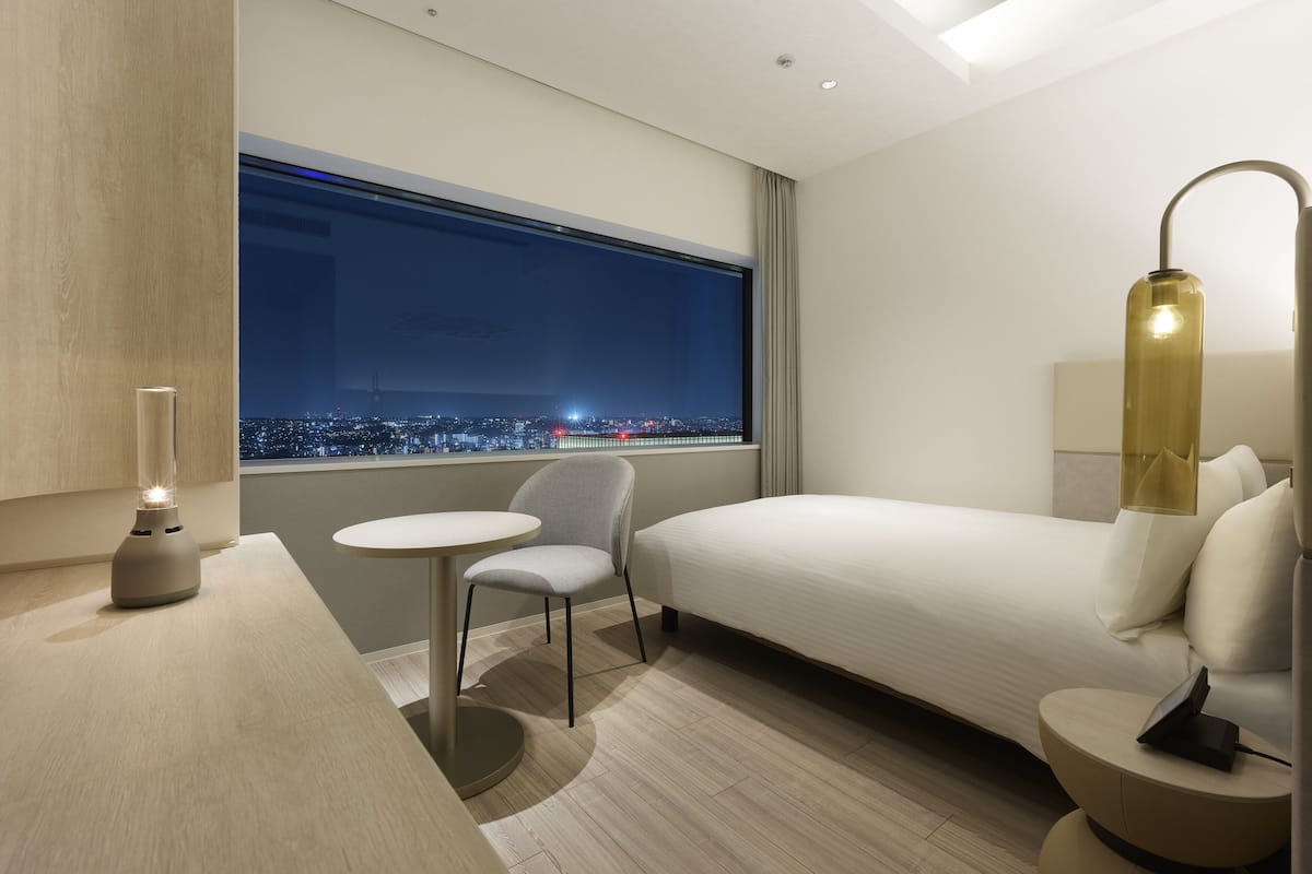 A modern hotel room with a bed, a small table, a chair, and a nightstand with a lamp.