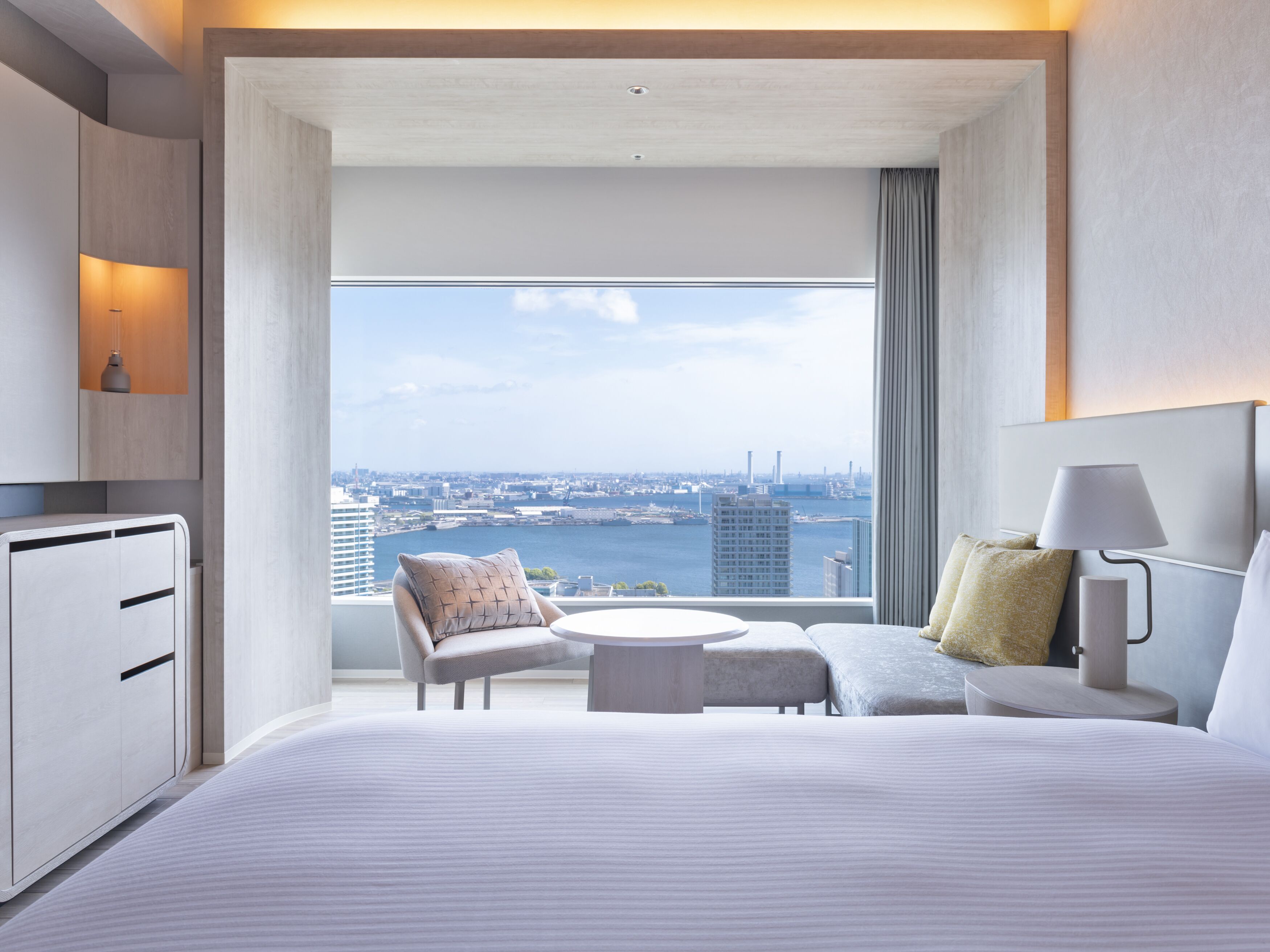 Ein modernes Hotelzimmer mit einem großen Bett, einer Sitzecke mit Tisch, einem Schreibtisch und Blick auf Stadt und Wasser.