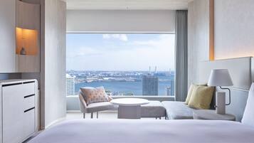 Ein modernes Hotelzimmer mit einem großen Bett, einer Sitzecke mit Tisch, einem Schreibtisch und Blick auf Stadt und Wasser.