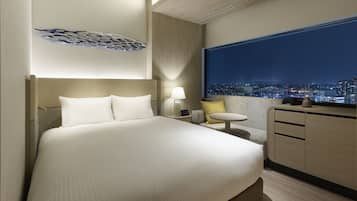 A modern hotel room with a large bed, a nightstand, a small table, and a view of a cityscape at night.