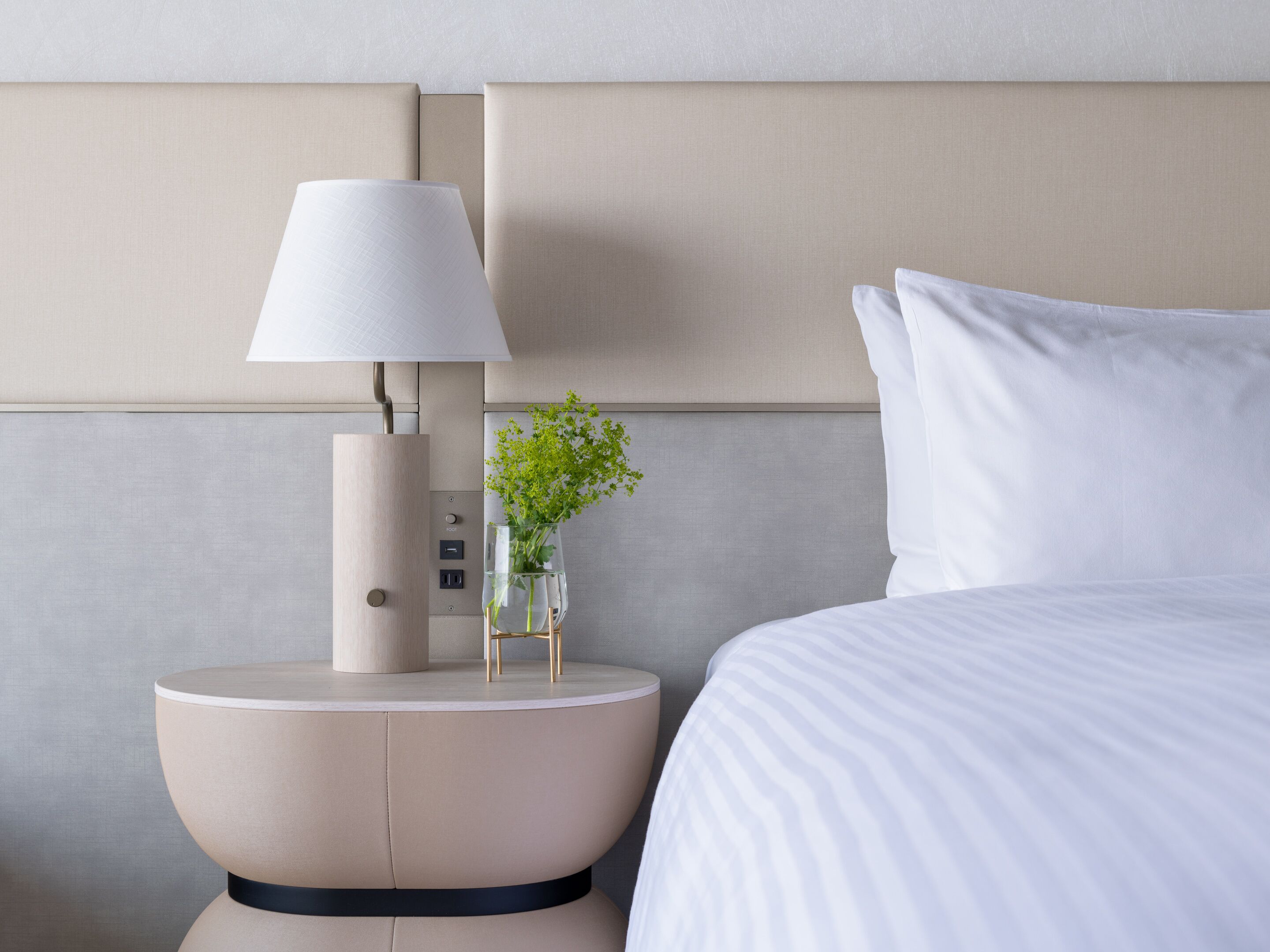 A modern hotel room with a bed, a bedside table with a lamp, and a vase with flowers.